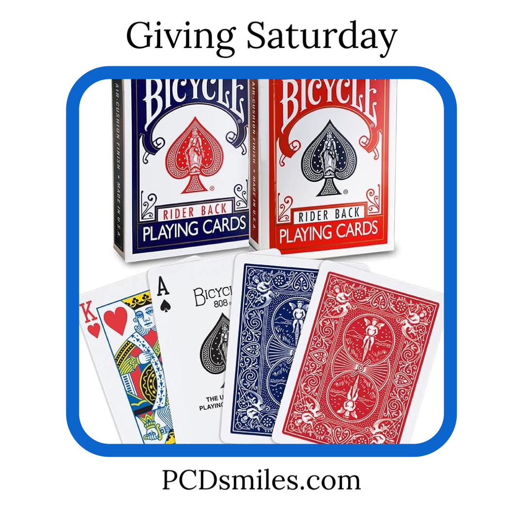 Giving Saturday Playing Cards Needed image is white background with black text reading Giving Saturday at the top, and black text reading PCDsmiles.com at bottom of graphic. Blue border around an image of a white background and two bicycle brand playing card boxes, one in red and one in blue. And four individual playing cards red king of hearts, black ace of spades, the back of a blue card, and the back of a red card.